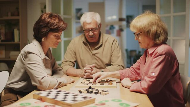 Medium shot of elderly friends playing checkers while sitting at table together at home