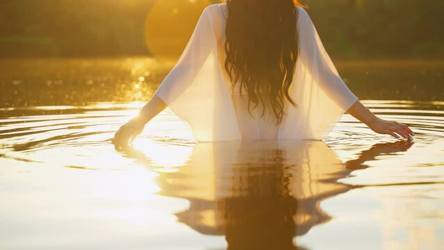 happy fantasy woman enjoys freedom magic warm sun light hot summer green nature. Girl elf stands in water river strokes lake, vintage wet white long silk dress back rear view. Sunset yellow flare 4k