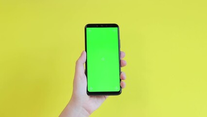 Chroma key mockup on smartphone in hand. Showcasing copy space for app or website design. A woman is holding a smartphone with a blank green screen on a yellow background