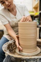 Fototapeta na wymiar Cropped view of blurred female potter making clay vase on pottery wheel in workshop at background