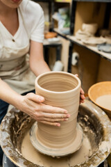 Fototapeta na wymiar Cropped view of female potter sculpting clay on pottery wheel in blurred ceramic workshop