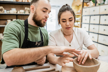 Obraz premium Bearded craftsman in apron shaping clay bowl and talking to girlfriend in ceramic workshop