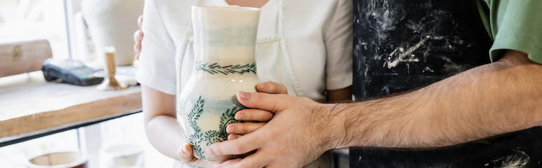 Naklejka premium Cropped view of couple of artisans in aprons holding clay vase in ceramic workshop, banner