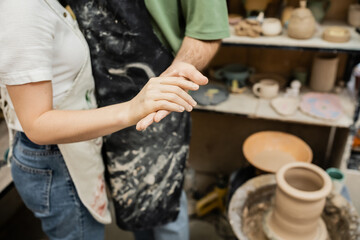 Cropped view of couple of sculptors in aprons holding hands near blurred  pottery wheel in workshop