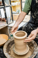 Cropped view of male artisan in apron shaping clay vase on pottery wheel near water in workshop