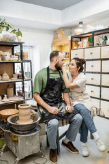 Craftswoman in apron hugging boyfriend and talking near clay on pottery wheel in workshop
