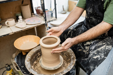 Fototapeta na wymiar Cropped view of potter in dirty apron shaping clay vase on pottery wheel near rack in workshop