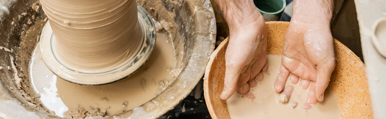 Fototapeta premium Top view of artisan working with water in bowl near clay and pottery wheel in ceramic studio, banner