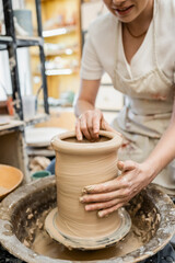Fototapeta na wymiar Partial view of female potter in blurred apron molding clay vase on pottery wheel in ceramic studio