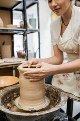 Fototapeta na wymiar Cropped view of female potter in apron creating clay vase on pottery wheel in blurred workshop