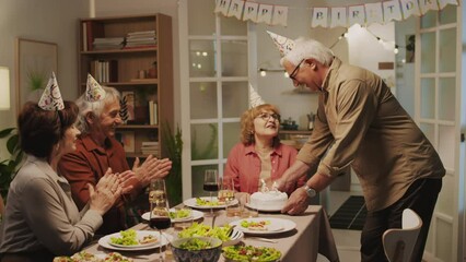 Long shot of positive senior husband bringing birthday cake to his wife and she blowing out candles in shape of number sixty five at home party