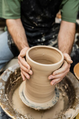 Cropped view of blurred potter in apron molding clay vase on pottery wheel in ceramic workshop