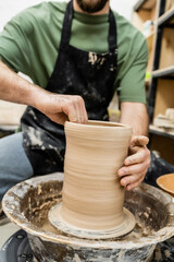 Fototapeta na wymiar Partial view of blurred artisan in apron shaping clay on pottery wheel in ceramic workshop