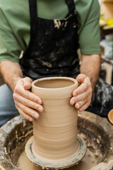 Fototapeta na wymiar Cropped view of blurred artisan in apron shaping clay vase on pottery wheel in ceramic studio