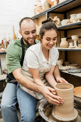 Cheerful craftsman hugging girlfriend and making clay vase together on pottery wheel in workshop