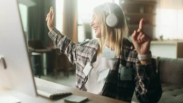 Portrait of cheerful blond mature woman freelancer with stickers listening to music in headphones ignore deadline at distance remote work on computer in home workplace No stress concept