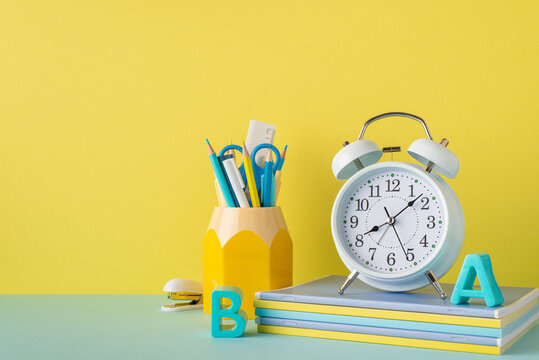 School supplies concept. Photo of stationery on blue desktop pencil holder alarm clock plastic alphabet letters stack of notebooks and stapler on yellow wall background