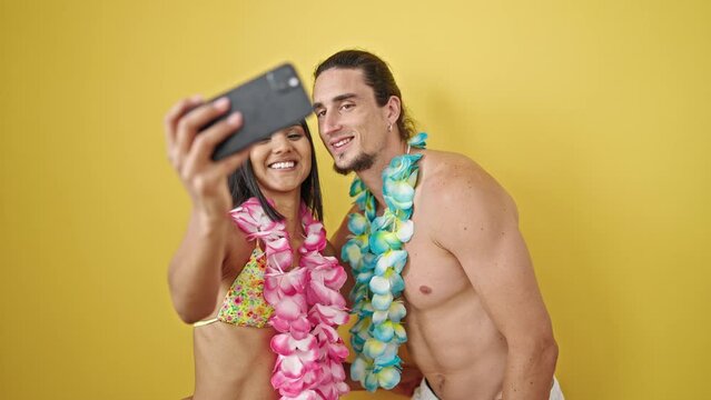 Man and woman tourist couple wearing hawaiian lei making selfie by smartphone over isolated yellow background