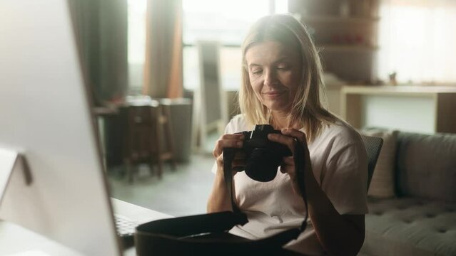 Portrait of pretty mature woman photographer hold digital camera looking at screen choosing photos for editing while sitting in front of computer at home workplace
