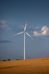 A wind turbine producing electricity standing in a field among cereals against a blue sky and clouds.