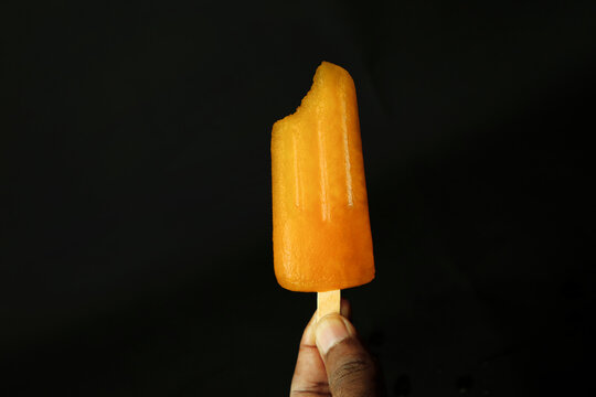 Orange Ice Cream On A Stick In Hand On A Black Background.