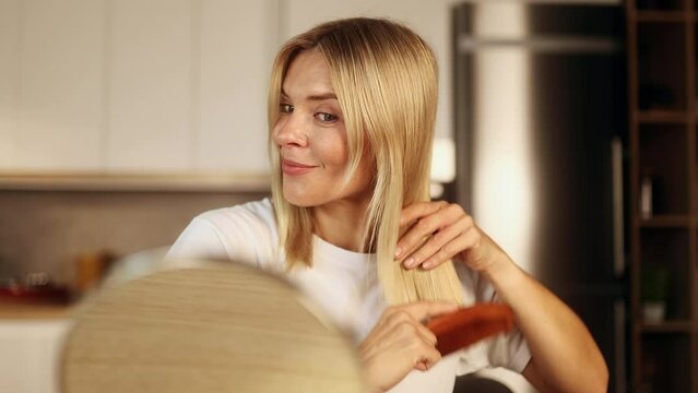 Portrait of pretty mature woman looking at mirror reflection and combing her blond hair with wooden brush in the morning at home Concept of hair growth trichology self care and daily routine