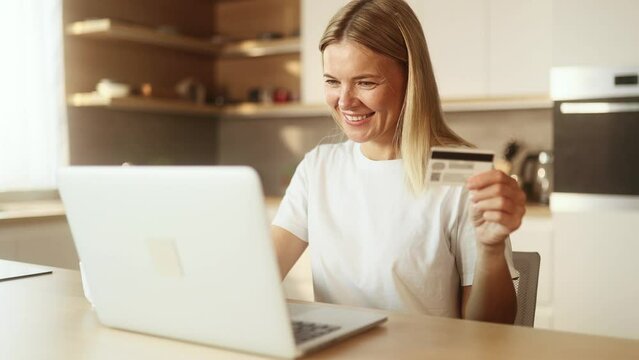 Portrait of charming mature blond woman paying with credit card on laptop at home Happy customer doing payments online shopping in internet store and receiving cash back indoors 