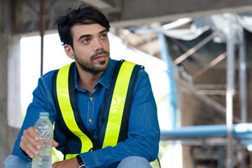 construction worker drinking water from bottle due to tired hard work and thirst, engineer wearing reflective vests take a rest sitting in area at building site