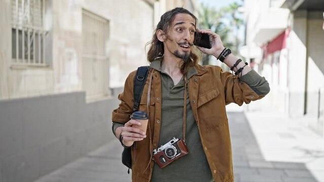 Young hispanic man tourist talking on smartphone holding coffee at street