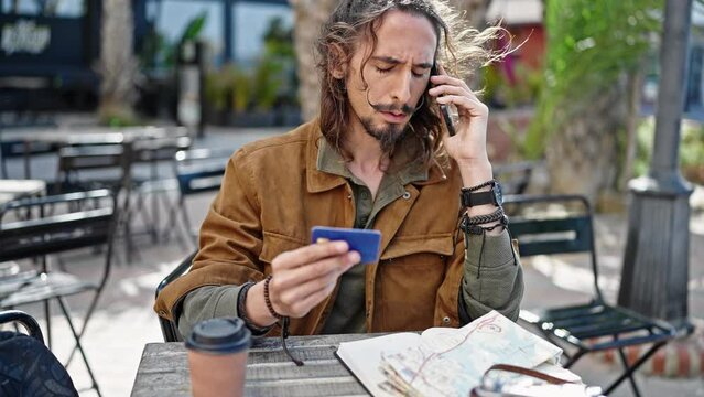 Young hispanic man tourist talking on smartphone holding credit card sitting on table at coffee shop terrace