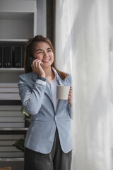 Portrait of young business woman with standing in office near the window and talking on mobile phone hand holding a cup of coffee