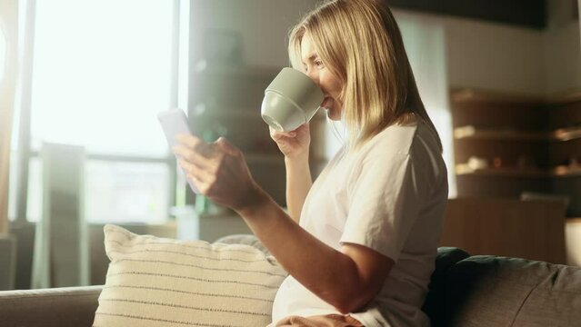 Portrait of charming blond mature woman with cup of hot coffee or tea hold smartphone scrolling watching social media indoors Happy female texting on her phone enjoying leisure time at home