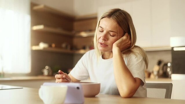 Boring exhausted mature woman watching online videos entertainment programs or news in social media on smartphone gadget while having breakfast in the morning at home