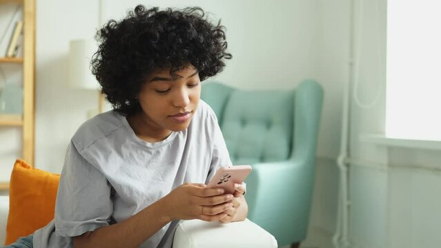 African girl holding smartphone touch screen typing scroll page at home. Woman with cell phone surfing internet using social media apps playing game. Shopping online Internet news cellphone addiction
