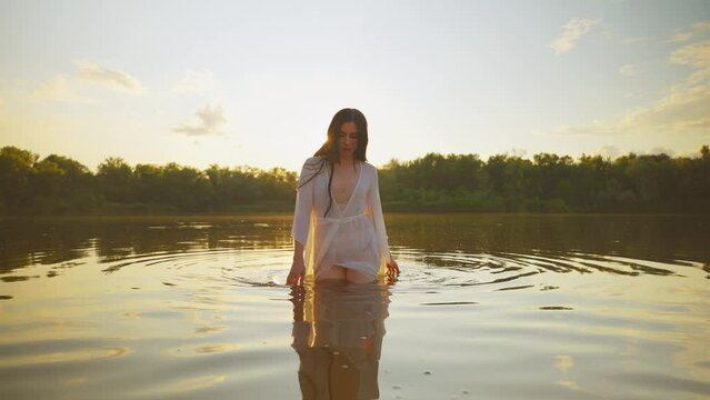 happy sexy fantasy woman emerges from water enjoys freedom magic sun light summer green nature. Girl river nymph hand strokes lake, vintage wet white long silk dress. Sunset yellow flare backlit. 4k