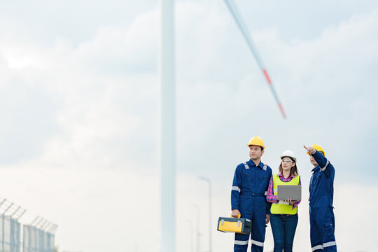 A team of 3 Asian male and female engineers with a mechanic manager point and look at the destination There is a wind turbine for the energy industry. holding via tablet tool box wear a helmet