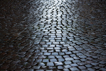 Cobblestone in a narrow street in Stresa, Italy. Wet shiny historic basalt ashlars or blocks reflecting blue sky and sunshine after rain. Old pavement background with typical surface and structure. - obrazy, fototapety, plakaty