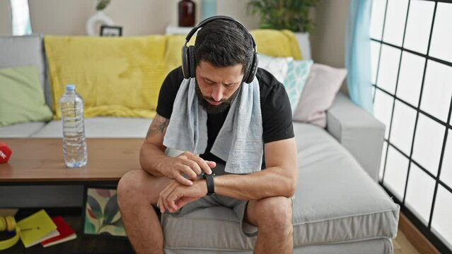 Young hispanic man sitting on sofa listening to music touching watch at home