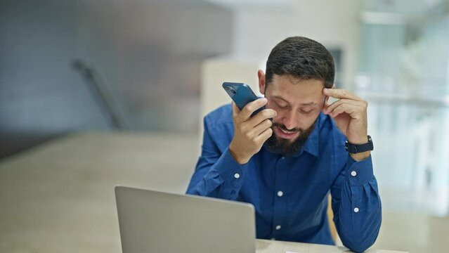 Young hispanic man business worker using laptop sending voice message by smartphone at the office