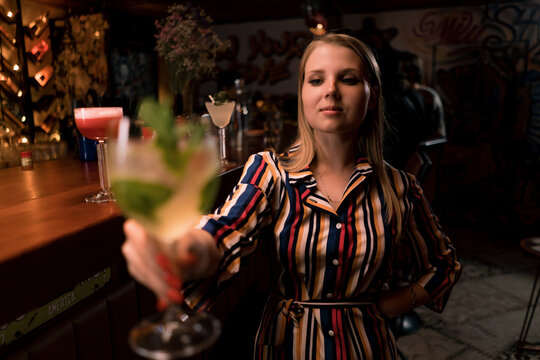 Young Cheerful Girl Holds Out A Mint Cocktail Towards The Camera To Make A Toast In A Club Bar