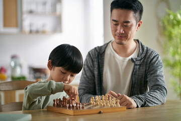 young asian father sitting at table playing chess with son at home