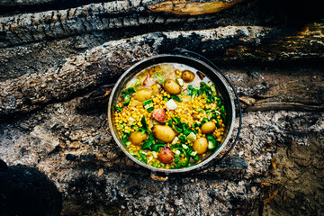 A vegetable stew of potatoes, green beans, and corn being cooked over hot coals on a campfire