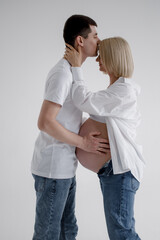 Family couple. A pregnant blond woman of Caucasian appearance is in bed with her husband, who kisses her on the forehead and gently touches her belly. They wear jeans and a white T-shirt.