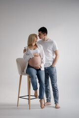 Family couple. A pregnant blond woman of Caucasian appearance is sitting on a chair, and her husband is standing on the side and gently kisses the woman. They stand on a white background. They wear je