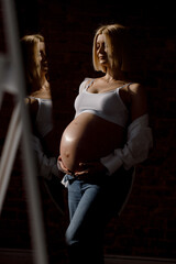 A beautiful pregnant woman of Caucasian appearance with white hair stands on a white background, touching her belly and looking away, dressed in a white top and jeans.