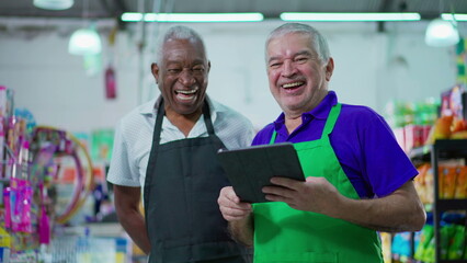Joyful diverse Brazilian senior staff workers of supermarket chain smiling at camera with table and uniforms. African American older employee and a caucasian person laughing together