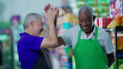 Happy diverse senior employees celebrating success with high-five standing in supermarket aisle. Caucasian manager engaging with workforce teamwork with African American colleague