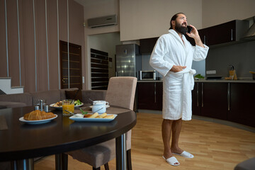 Bearded male in a bathrobe communicates on a mobile phone - obrazy, fototapety, plakaty