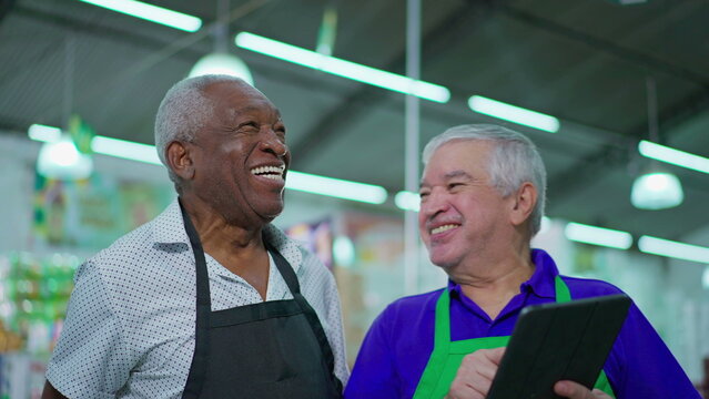 Happy senior colleagues of supermarket laughing and smiling, candid authentic joyful interaction between older diverse staff men while holding tablet device