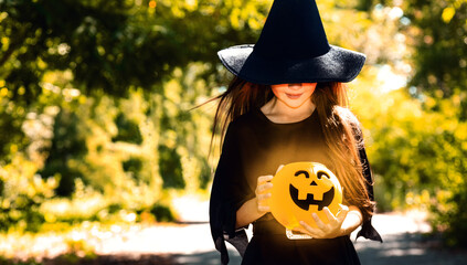 Halloween. Little girl in witch costume r with lantern pumpkin. Child in black hat plays in horror stories outside